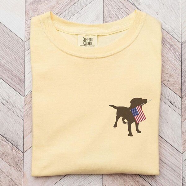 Butter yellow T-shirt laid on a wood floor with a chocolate lab carrying an American flag on the left chest