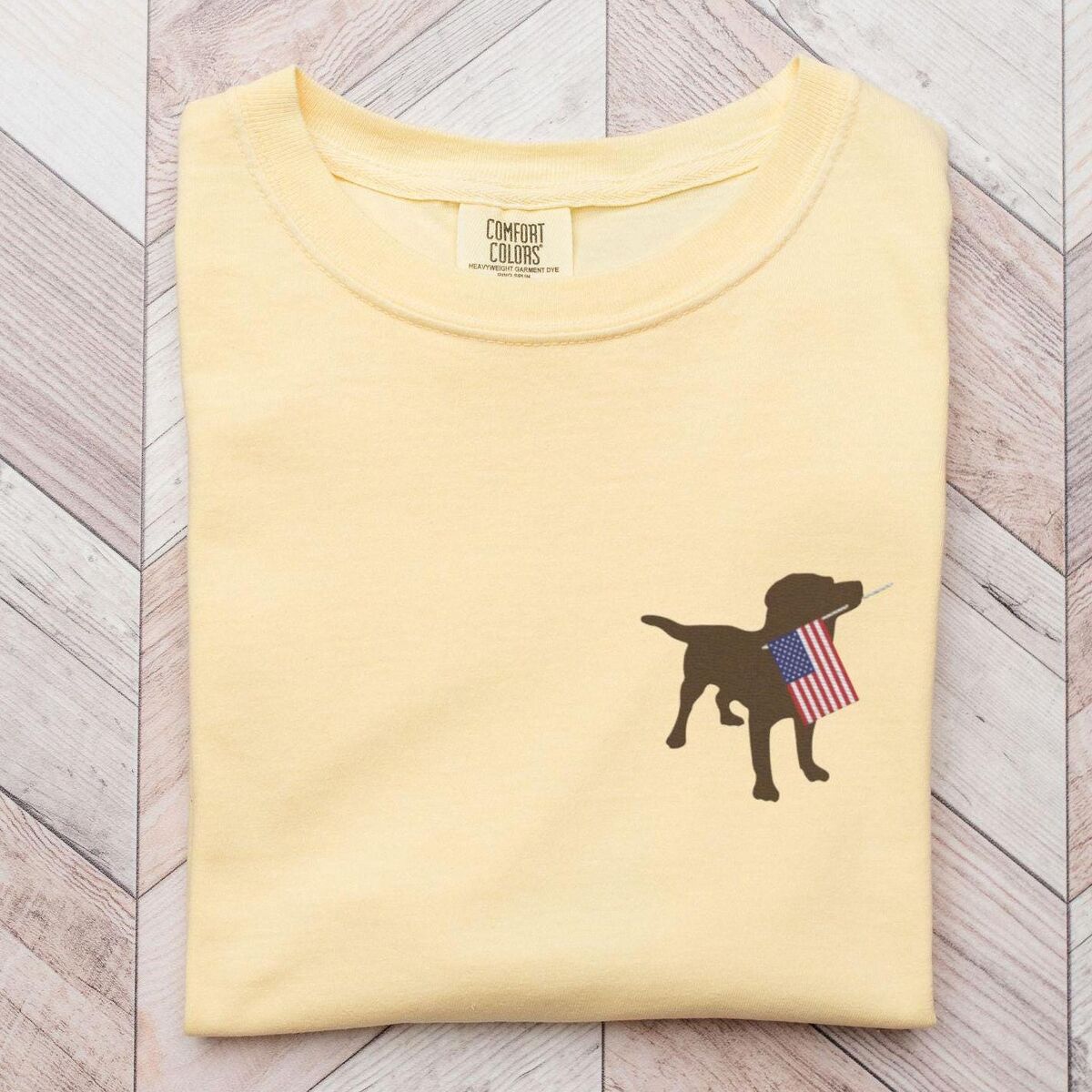 Chocolate Lab American Flag Shirt - Butter Butter yellow T-shirt laid on a wood floor with a chocolate lab carrying an American flag on the left chest