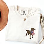 Ivory T-shirt folded beside a hat with a chocolate lab silhouette carrying an American flag on the left chest