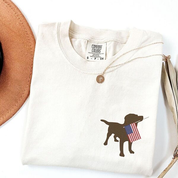 Ivory T-shirt folded beside a hat with a chocolate lab silhouette carrying an American flag on the left chest