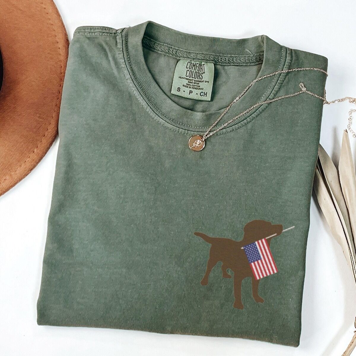 Chocolate Lab American Flag Shirt – Moss Moss T-shirt folded with a necklace, a brown Labrador holding a stars-and-stripes flag stitched on the left chest