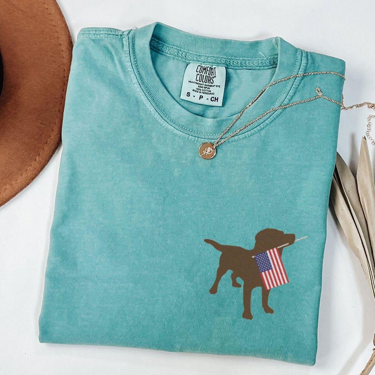 Chocolate Lab American Flag Shirt – Seafoam Seafoam T-shirt folded with a necklace, a chocolate lab carrying a red, white and blue flag on the left chest