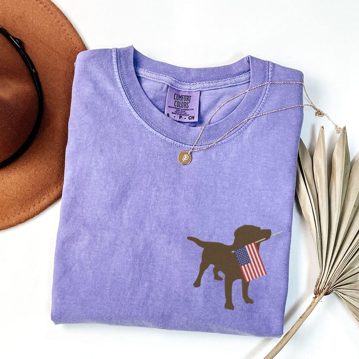 Chocolate Lab American Flag Shirt – Violet Violet T-shirt folded beside a hat and dried palm leaf, embroidered with a chocolate lab carrying a USA flag
