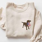 Sand crewneck sweatshirt laid flat with a rolled sleeve, a small chocolate lab silhouette carrying a USA flag on the left chest