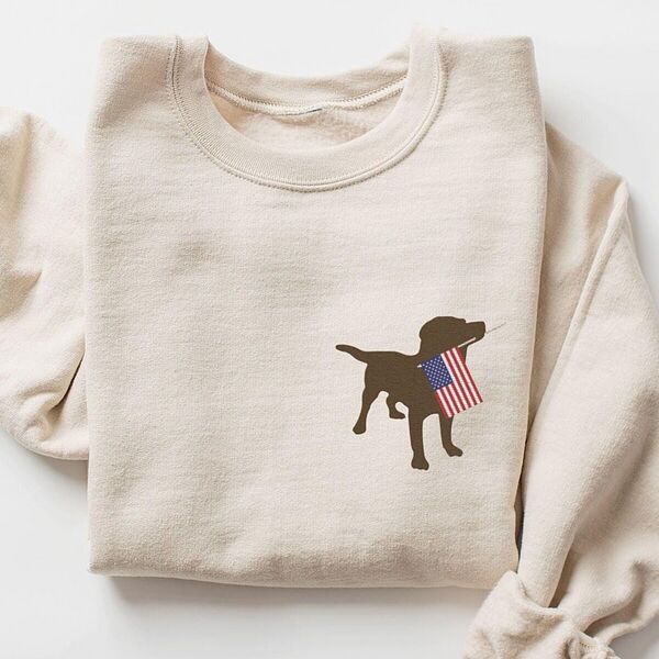 Sand crewneck sweatshirt laid flat with a rolled sleeve, a small chocolate lab silhouette carrying a USA flag on the left chest