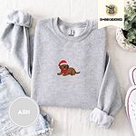 Ash crewneck sweatshirt folded with denim shorts, white sneakers and a potted plant, embroidered with a chocolate lab puppy in a Santa hat and red scarf in the center of the chest