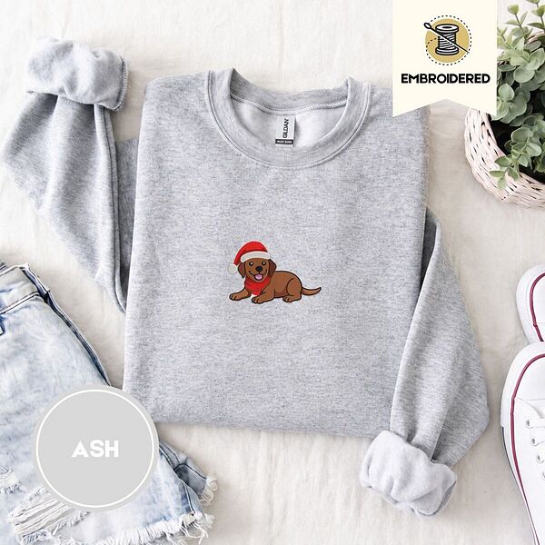 Ash crewneck sweatshirt folded with denim shorts, white sneakers and a potted plant, embroidered with a chocolate lab puppy in a Santa hat and red scarf in the center of the chest