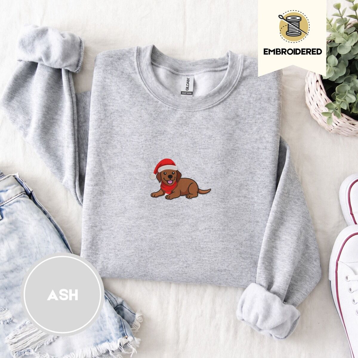 Chocolate Lab Christmas Sweatshirt – Ash Ash crewneck sweatshirt folded with denim shorts, white sneakers and a potted plant, embroidered with a chocolate lab puppy in a Santa hat and red scarf in the center of the chest