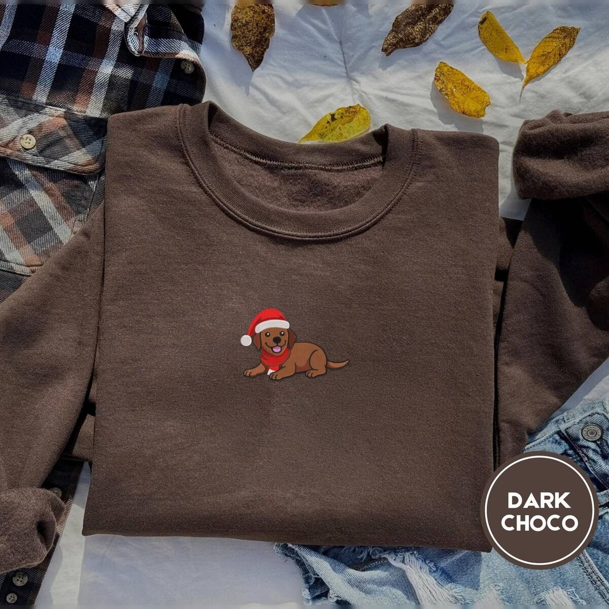 Chocolate Lab Christmas Sweatshirt – Dark Chocolate Dark Chocolate crewneck sweatshirt on a plaid flannel shirt with yellow autumn leaves and denim shorts, embroidered with a chocolate lab puppy in a Santa hat and red scarf in the center of the chest