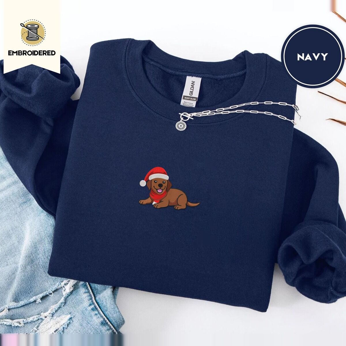 Chocolate Lab Christmas Sweatshirt – Navy Navy crewneck sweatshirt folded with ripped jeans and a silver chain necklace, embroidered with a chocolate lab puppy in a Santa hat and red scarf in the center of the chest