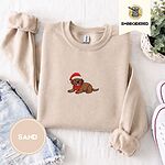 Sand crewneck sweatshirt folded with denim shorts, white sneakers and a potted plant, embroidered with a chocolate lab puppy in a Santa hat and red scarf in the center of the chest