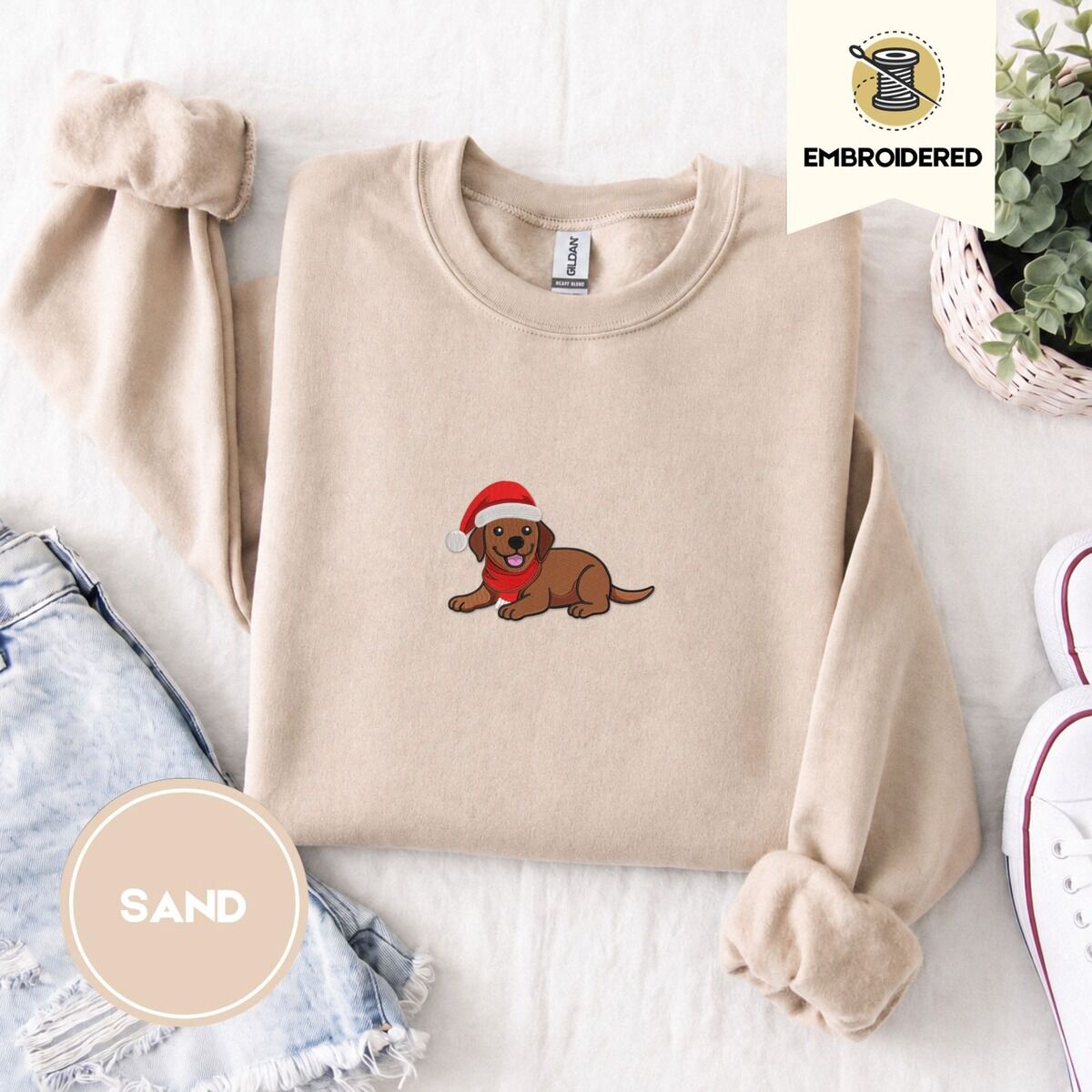 Chocolate Lab Christmas Sweatshirt – Sand Sand crewneck sweatshirt folded with denim shorts, white sneakers and a potted plant, embroidered with a chocolate lab puppy in a Santa hat and red scarf in the center of the chest