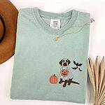 Bay T-shirt laid flat with a necklace and dried palm leaves, embroidered with a brown Labrador trick-or-treating in a ghost sheet