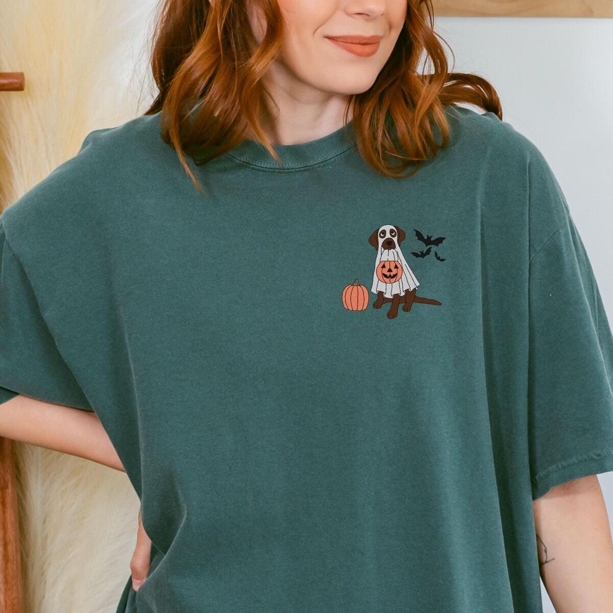 Chocolate Lab Ghost Shirt – Blue Spruce Smiling woman in a Blue Spruce T-shirt embroidered with a chocolate lab trick-or-treating in a ghost sheet on the left chest