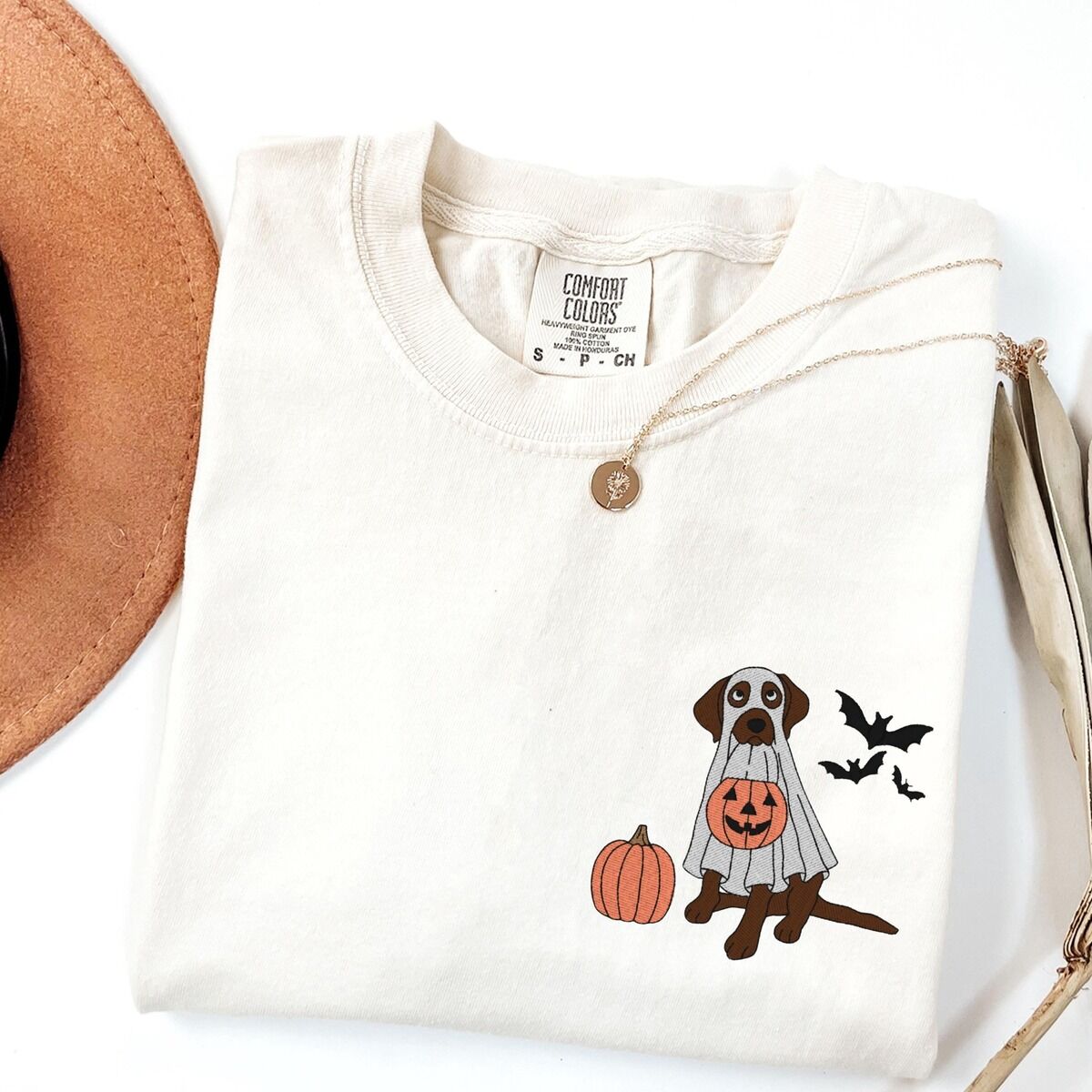 Chocolate Lab Ghost Shirt – Ivory Ivory T-shirt laid flat with a necklace, embroidered with a chocolate lab in a white ghost costume, a pumpkin and three bats