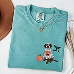 Seafoam T-shirt laid flat with a necklace, embroidered with a chocolate Labrador in a ghost costume beside a pumpkin and three bats
