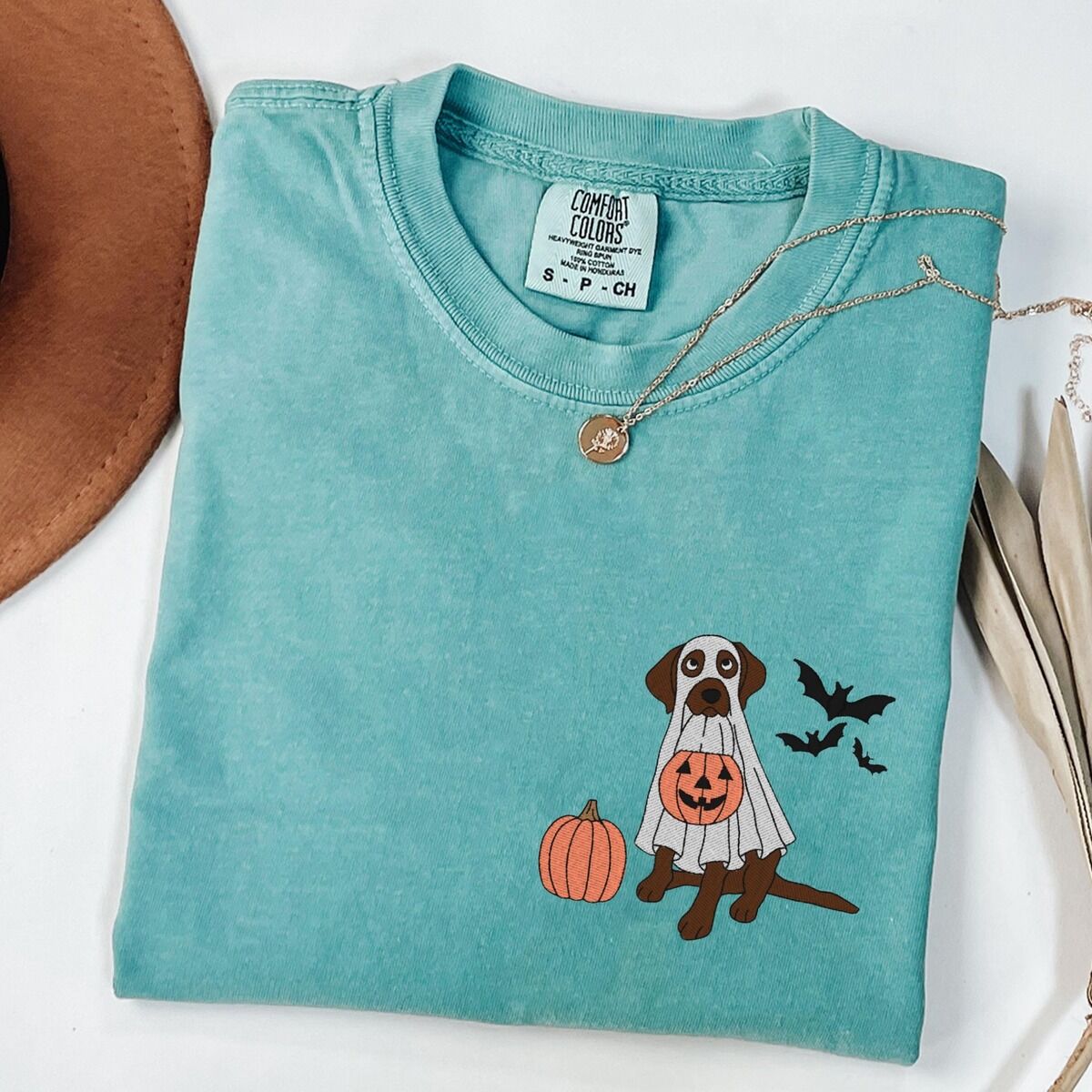 Chocolate Lab Ghost Shirt – Seafoam Seafoam T-shirt laid flat with a necklace, embroidered with a chocolate Labrador in a ghost costume beside a pumpkin and three bats