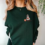 Woman in a Forest Green crewneck sweatshirt embroidered with a small chocolate lab in a ghost costume, a pumpkin and bats on the left chest