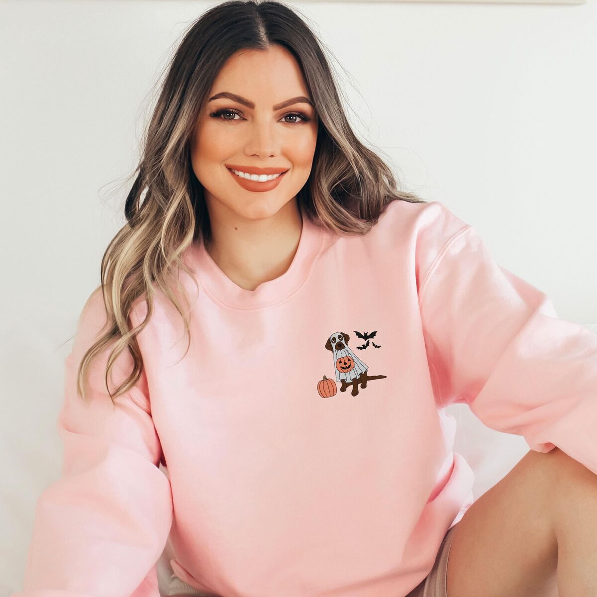 Chocolate Lab Ghost Sweatshirt - Light Pink Smiling woman in a Light Pink crewneck sweatshirt embroidered with a small chocolate Labrador in a ghost sheet holding a jack-o'-lantern bucket