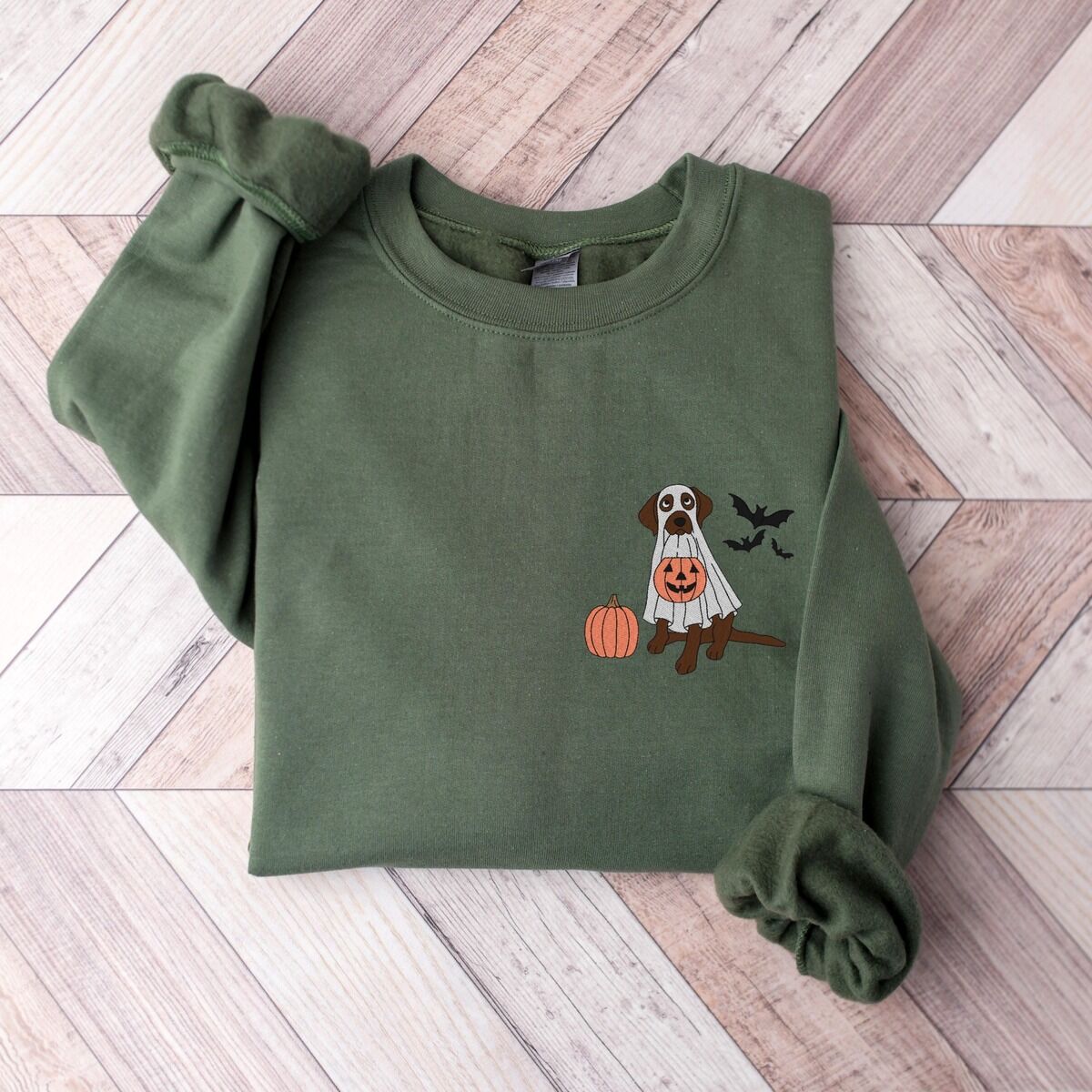 Chocolate Lab Ghost Sweatshirt – Military Green Chocolate lab ghost sweatshirt in Military Green laid flat on a herringbone wood floor, embroidered with a brown Labrador in a ghost sheet holding a jack-o'-lantern bucket beside a pumpkin and three bats