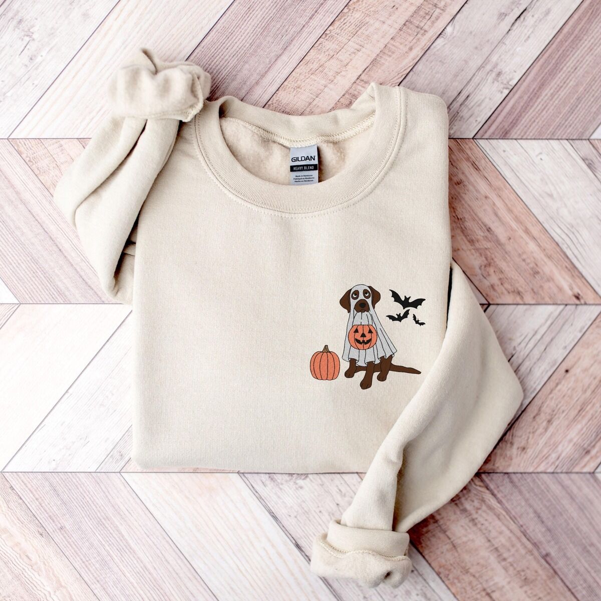 Chocolate Lab Ghost Sweatshirt – Sand Sand crewneck sweatshirt on a wood floor, embroidered with a brown lab under a ghost sheet with a jack-o'-lantern bucket on the left chest