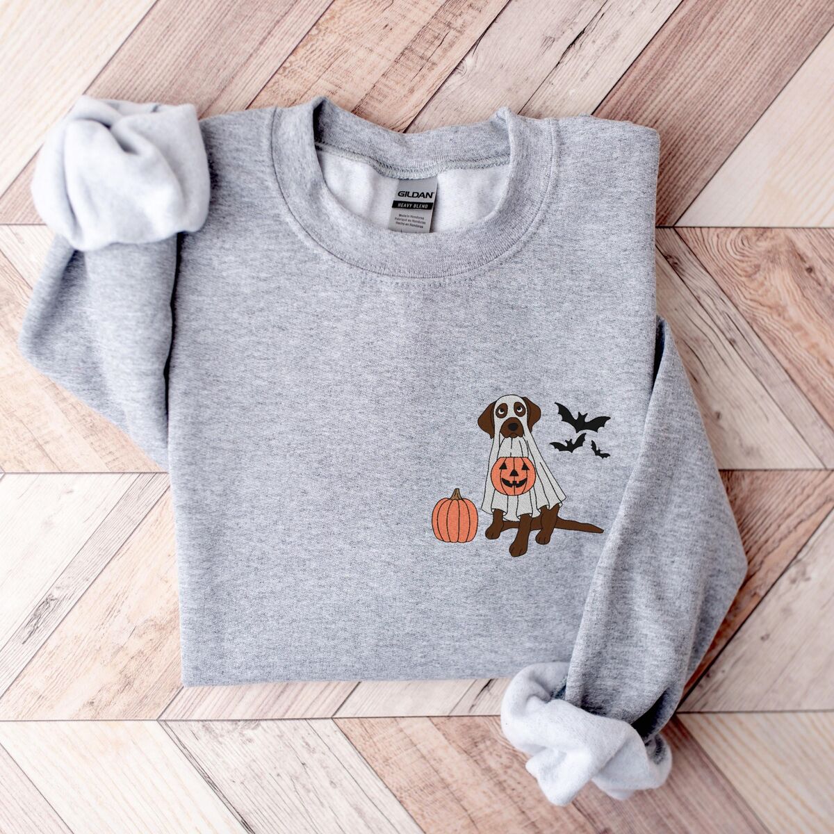 Chocolate Lab Ghost Sweatshirt - Sport Grey Sport Grey crewneck sweatshirt on a herringbone wood floor, embroidered with a brown lab in a ghost sheet holding a jack-o'-lantern bucket beside three bats