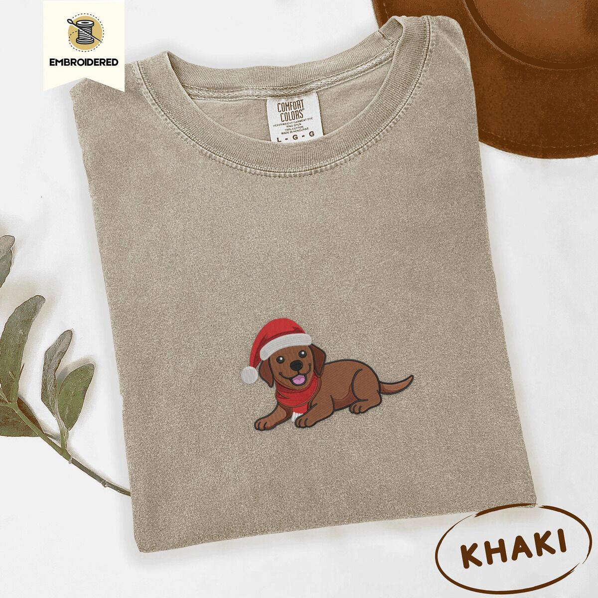 Chocolate Lab Santa Hat Shirt – Khaki Khaki Comfort Colors T-shirt embroidered with a chocolate Labrador puppy lying down in a red Santa hat and a red knit scarf