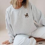 Woman sitting in an Ash crewneck sweatshirt embroidered with a small chocolate brown Labrador silhouette in a blue collar and gold bone tag on the left chest