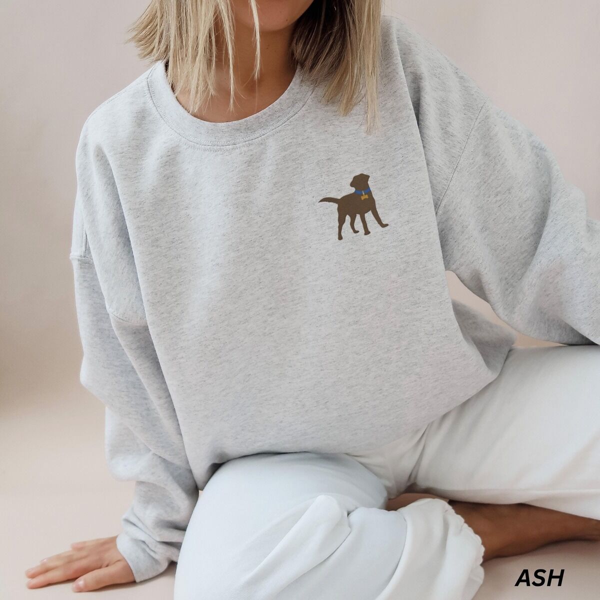 Chocolate Lab Silhouette Sweatshirt – Ash Woman sitting in an Ash crewneck sweatshirt embroidered with a small chocolate brown Labrador silhouette in a blue collar and gold bone tag on the left chest