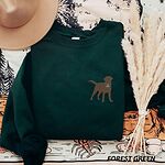 Forest Green crewneck sweatshirt laid flat with a hat and pampas grass, embroidered with a small chocolate brown Labrador silhouette in a blue collar and gold bone tag on the left chest