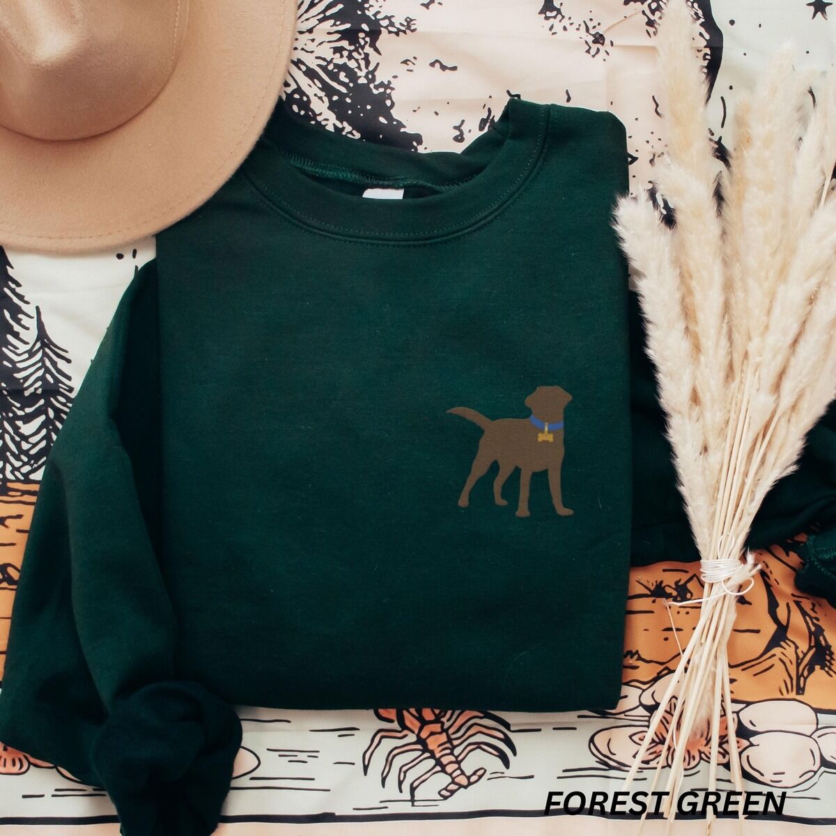 Chocolate Lab Silhouette Sweatshirt – Forest Green Forest Green crewneck sweatshirt laid flat with a hat and pampas grass, embroidered with a small chocolate brown Labrador silhouette in a blue collar and gold bone tag on the left chest
