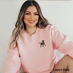 Smiling woman in a Light Pink crewneck sweatshirt embroidered with a small chocolate brown Labrador silhouette in a blue collar and gold bone tag on the left chest