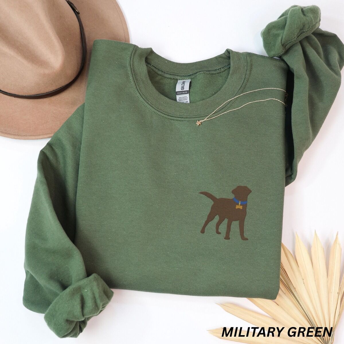 Chocolate Lab Silhouette Sweatshirt – Military Green Military Green crewneck sweatshirt laid flat with a hat and a necklace, embroidered with a small chocolate brown Labrador silhouette in a blue collar and gold bone tag on the left chest