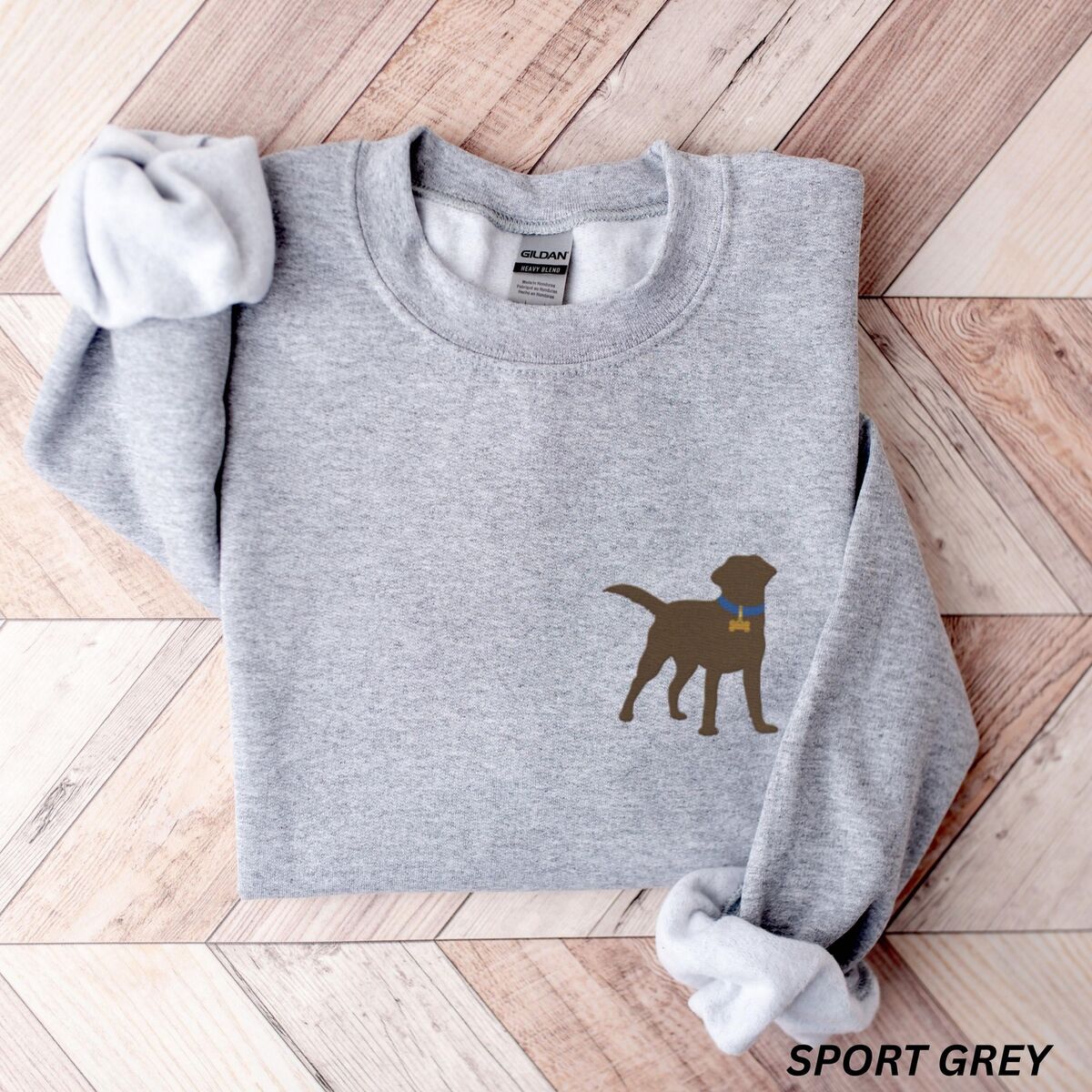 Chocolate Lab Silhouette Sweatshirt - Sport Grey Sport Grey crewneck sweatshirt laid flat on herringbone wood, embroidered with a small chocolate brown Labrador silhouette in a blue collar and gold bone tag on the left chest