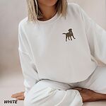 Woman sitting in a White crewneck sweatshirt embroidered with a small chocolate brown Labrador silhouette in a blue collar and gold bone tag on the left chest