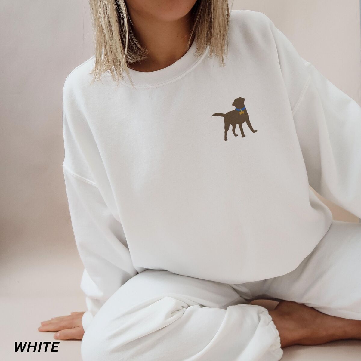 Chocolate Lab Silhouette Sweatshirt - White Woman sitting in a White crewneck sweatshirt embroidered with a small chocolate brown Labrador silhouette in a blue collar and gold bone tag on the left chest