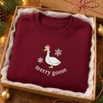Maroon Christmas goose crewneck folded in a gift box with white tissue paper and gold ornaments