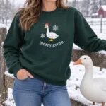 Woman wearing the Forest Green merry goose sweatshirt by a snowy farm fence with a white goose