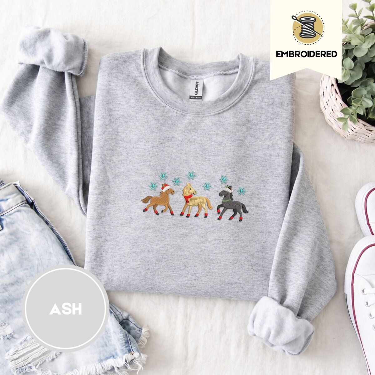 Christmas Horse Sweatshirt – Ash Ash crewneck sweatshirt folded with denim shorts, white sneakers and a potted plant, embroidered with three horses in Santa hats, a red scarf and red leg wraps under turquoise snowflakes