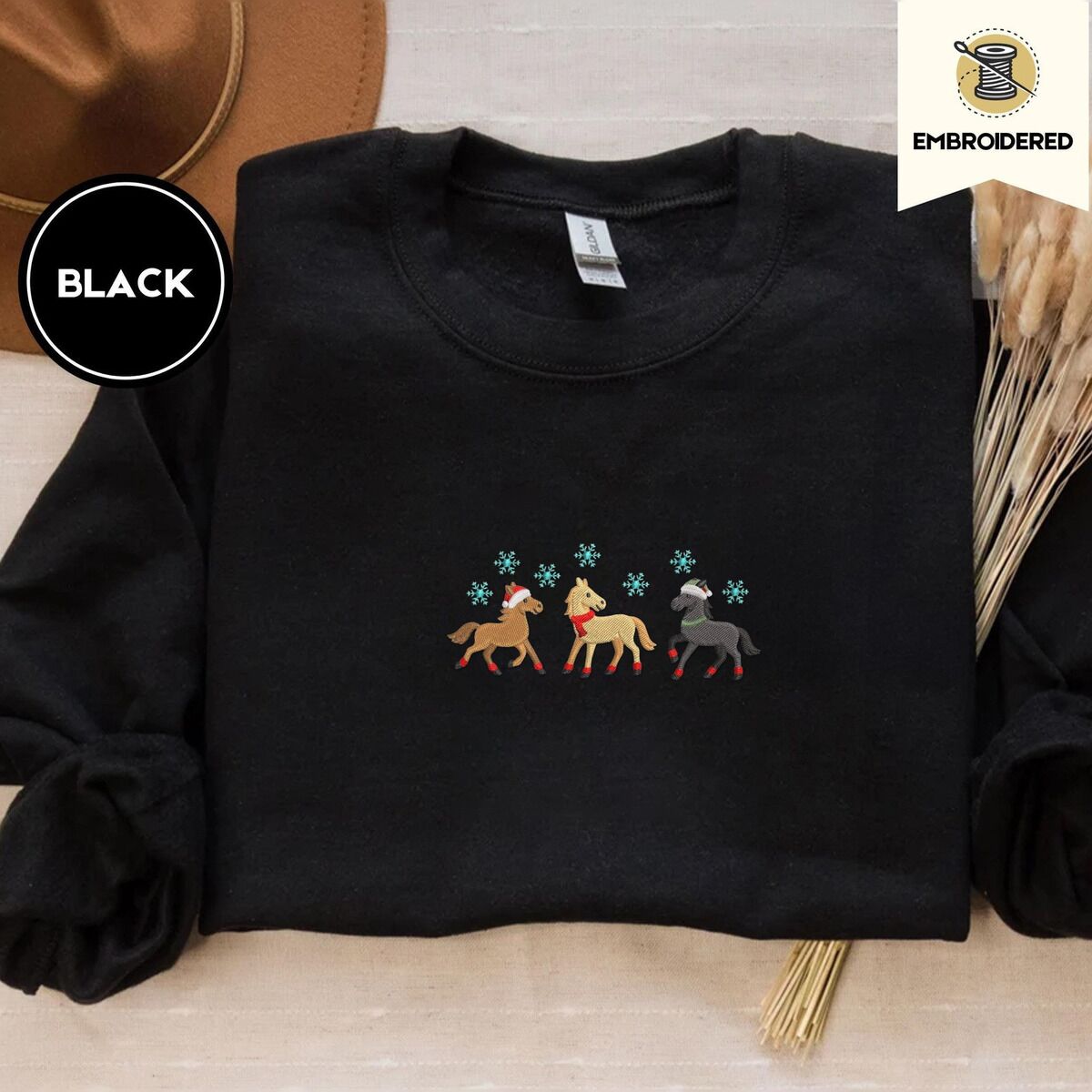 Christmas Horse Sweatshirt – Black Black crewneck sweatshirt with a brown felt hat and dried bunny tail grass, embroidered with three horses in Santa hats, a red scarf and red leg wraps under turquoise snowflakes