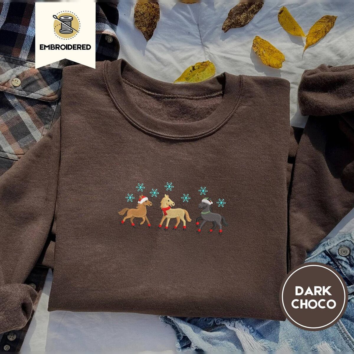 Christmas Horse Sweatshirt – Dark Chocolate Dark Chocolate crewneck sweatshirt on a plaid flannel shirt with yellow autumn leaves and denim shorts, embroidered with three horses in Santa hats, a red scarf and red leg wraps under turquoise snowflakes