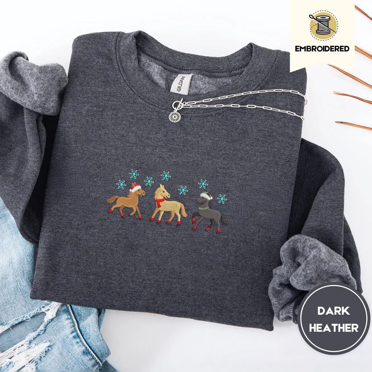 Christmas Horse Sweatshirt – Dark Heather Dark Heather crewneck sweatshirt folded with ripped jeans and a silver chain necklace, embroidered with three horses in Santa hats, a red scarf and red leg wraps under turquoise snowflakes