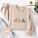 Sand crewneck sweatshirt folded with denim shorts, white sneakers and a potted plant, embroidered with three horses in Santa hats, a red scarf and red leg wraps under turquoise snowflakes