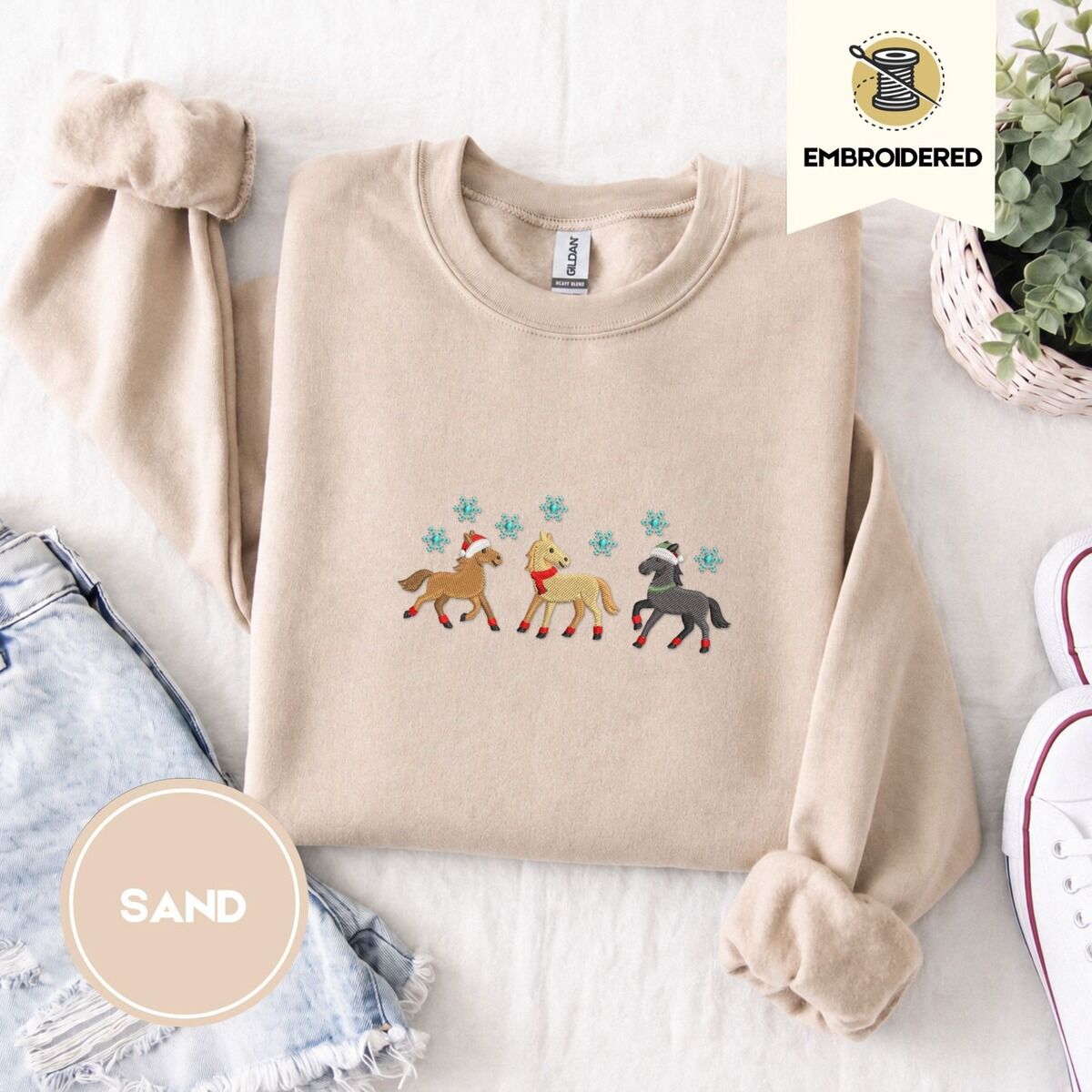 Christmas Horse Sweatshirt – Sand Sand crewneck sweatshirt folded with denim shorts, white sneakers and a potted plant, embroidered with three horses in Santa hats, a red scarf and red leg wraps under turquoise snowflakes