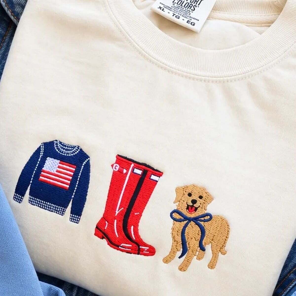 Coastal Icons Sweatshirt – Ivory Ivory Comfort Colors crewneck sweatshirt embroidered with a navy flag sweater, red rain boots and a golden retriever in a navy bow