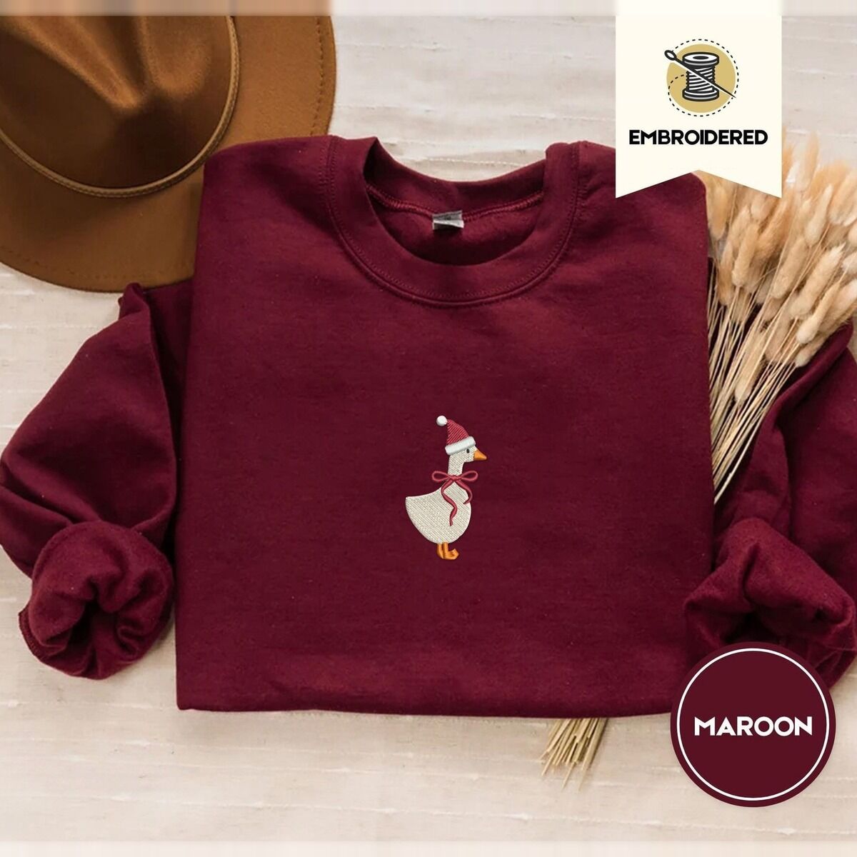 Santa Goose Sweatshirt – Maroon Maroon crewneck sweatshirt with a brown felt hat and dried bunny tail grass, embroidered with a white goose in a Santa hat and a red ribbon bow in the center of the chest