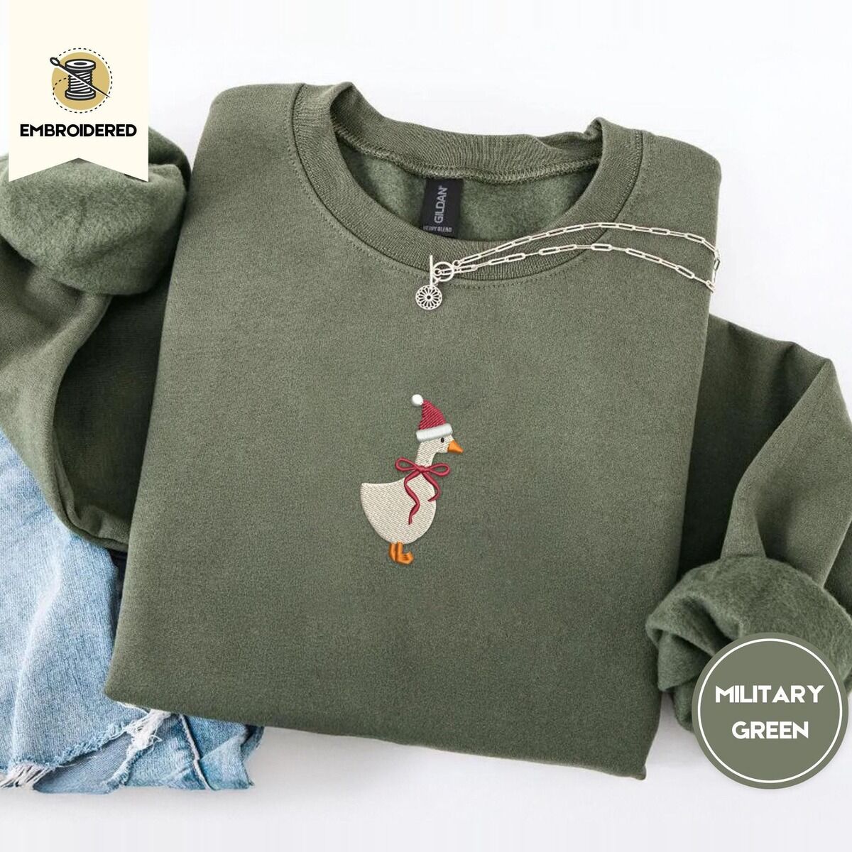 Santa Goose Sweatshirt – Military Green Military Green crewneck sweatshirt folded with light jeans and a silver chain necklace, embroidered with a white goose in a Santa hat and a red ribbon bow in the center of the chest