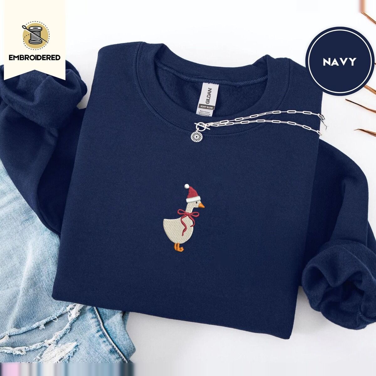 Santa Goose Sweatshirt – Navy Navy crewneck sweatshirt folded with ripped jeans and a silver chain necklace, embroidered with a white goose in a Santa hat and a red ribbon bow in the center of the chest