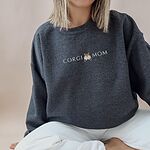 Woman sitting cross-legged in a Dark Heather crewneck sweatshirt embroidered with CORGI MOM and a corgi butt