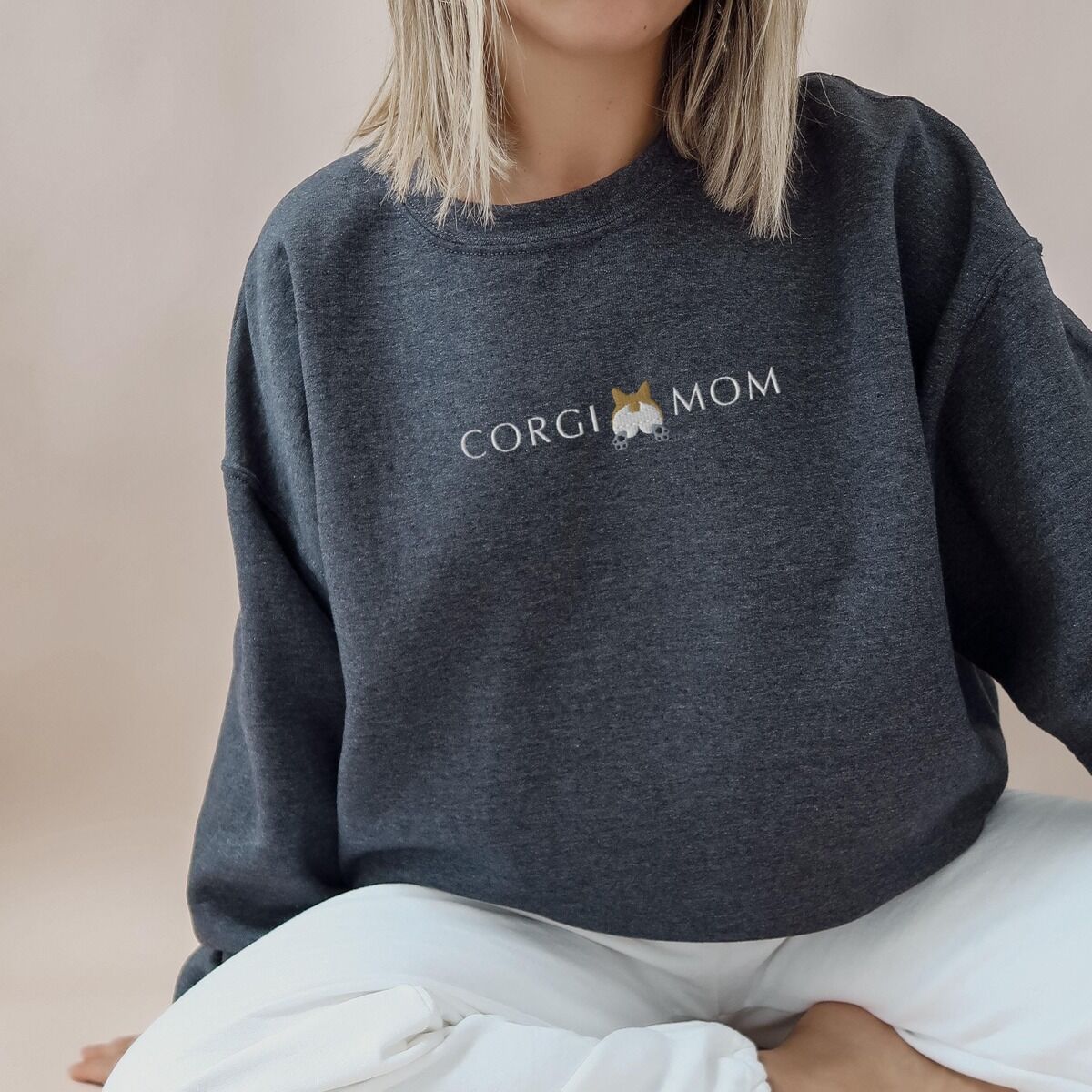 Corgi Butt Sweatshirt – Dark Heather Woman sitting cross-legged in a Dark Heather crewneck sweatshirt embroidered with CORGI MOM and a corgi butt